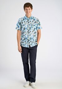 Korte mouwen shirt met een lichtblauwe achtergrond met een bloemen- en bladerenpatroon. Gecombineerd met donkere broeken en witte sneakers.