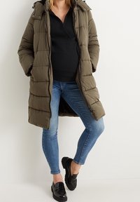 Olivengrøn dunjakke med hætte, quiltet design og lynlåslukning, kombineret med mørk top, tætsiddende blå jeans og sorte loafers.