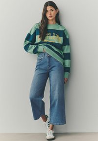 Sweatshirt rayé vert et marine avec le texte "Portland Oregon", associé à un jean en denim taille large bleu clair et des baskets blanches avec des accents verts.