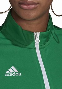 Zelená bunda na zip z texturované látky s vysokým límcem. Má bílý zip a doplňky, na hrudi je bílé logo Adidas.