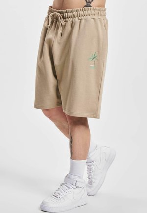Shorts en coton beige avec une taille élastique et un cordon de serrage. Présente un petit graphique d'un palmier vert sur la cuisse gauche. Porté avec des baskets blanches.