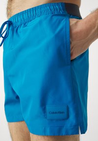 Costumi da bagno blu realizzati in tessuto leggero, dotati di una vita elastica con cordino, tasche laterali e una patch nera con logo.