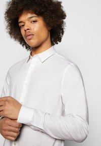 Camicia bianca con bottoni realizzata in tessuto liscio; presenta un colletto classico, maniche lunghe e un logo sottile sul petto sinistro.