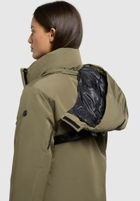 Oliven grøn parka-jakke med høj krave, der har et sort inderfor og justerbar hætte, der er sikret med velcro og hardware-detaljer.