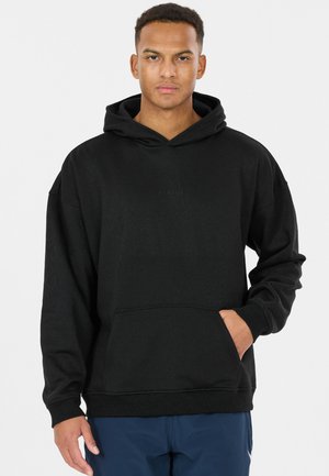 Man draagt een zwarte capuchonsweater met een voorzak en donkerblauwe broek, staand tegen een effen lichte achtergrond.