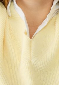 Sweter w serek z miękkiej, teksturowanej żółtej dzianiny z subtelnymi detalami w prążki, nałożony na białą koszulę z kołnierzykiem.