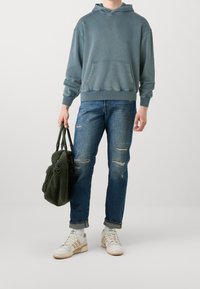 Felpa blu foderata in pile con tasca a marsupio, jeans blu strappati, sneakers beige con accenti verdi e una borsa tote di pelliccia verde.