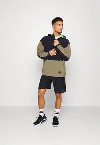 Ανοικτό μπουφάν με κουκούλα σε ναυτικό και ελιά, με λογότυπο ρόμβου· συνδυασμένο με μαύρα σορτς, κάλτσες λευκές με κόκκινες ρίγες και μαύρα αθλητικά παπούτσια.