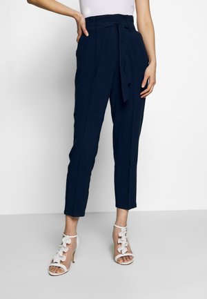 Kobieta w granatowych, wysokich, zwężanych spodniach z materiałowym paskiem, w połączeniu z białymi sandałami na obcasie ozdobionymi kwiatowymi detalami.