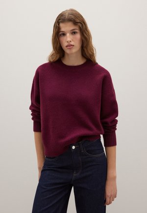 Suéter burdeos con cuello redondo, mangas largas y puños acanalados, combinado con jeans de un azul oscuro. La tela parece suave y con textura.