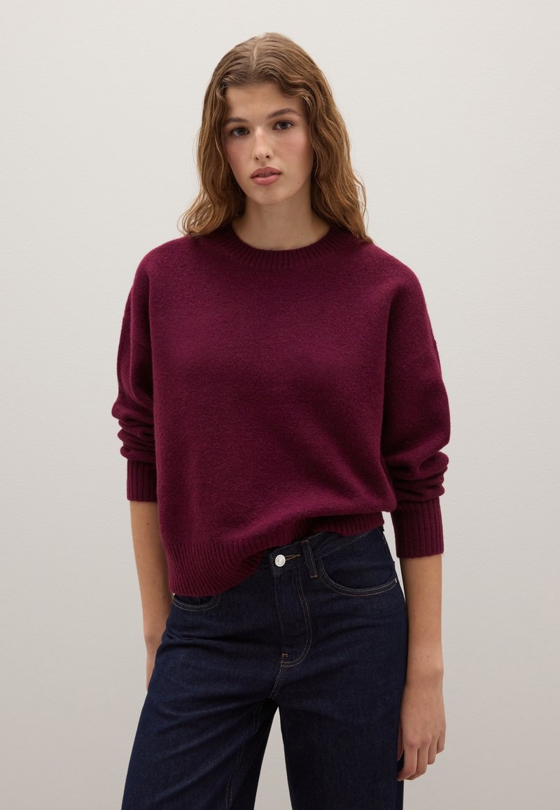 Bordeauxfarbener Pullover mit runder Halslinie, langen Ärmeln und gerippten Bündchen, kombiniert mit dunkelblauen Jeans. Der Stoff wirkt weich und strukturiert.