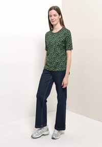 Femme debout, les yeux fermés, portant un haut vert à motifs à manches courtes, un pantalon bleu foncé à jambes larges et des baskets argentées.