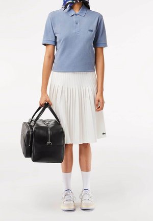 Personne portant un polo bleu et une jupe plissée blanche, tenant un sac de voyage en cuir noir, avec des chaussettes blanches et des baskets, debout sur un fond blanc.