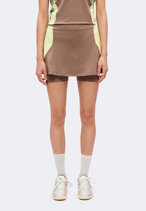 Personne portant une tenue short-jupe marron avec des panneaux latéraux jaunes, des chaussettes blanches aux chevilles et des chaussures de sport blanches sur un fond uni.