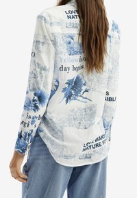 Langarmshirt in Weiß und Blau mit floralem Muster und Textdetails. Leichter Stoff mit lockerer Passform und abgerundetem Saum.