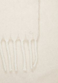 Foulard doux et léger de couleur crème, avec des bords frangés. Confectionné en matériau fin et texturé avec un motif subtil. Présente une petite étiquette de marque sur un côté.