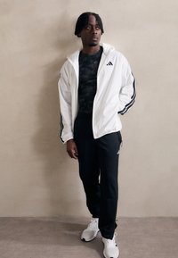 Casaco branco com fecho e riscas pretas, camisa justa preta, calças pretas e sapatilhas brancas. O modelo está de pé num fundo neutro.