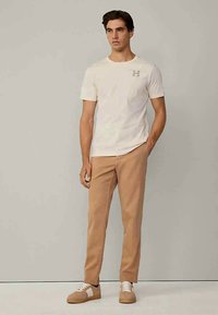 Man som bär en vit T-shirt med kort ärm och ett "H"-logo, beige byxor och beige sneakers, står med en hand i fickan framför en enkel bakgrund.