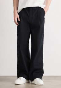 Pantaloni negri cu croială largă, din bumbac, cu faț�ă plată, buzunare laterale și o textură netedă. Purtați cu adidași albi.