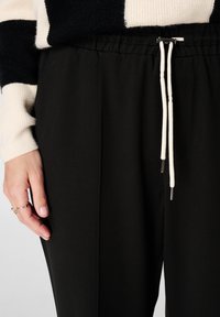 Pantalon de jogging noir avec taille élastique et cordon blanc, doté d'embouts métalliques argentés. Texture lisse et coupe décontractée.