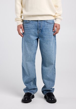 Jean en denim bleu clair avec une coupe décontractée, un design à cinq poches et une texture légèrement délavée. Associé à des chaussures noires.