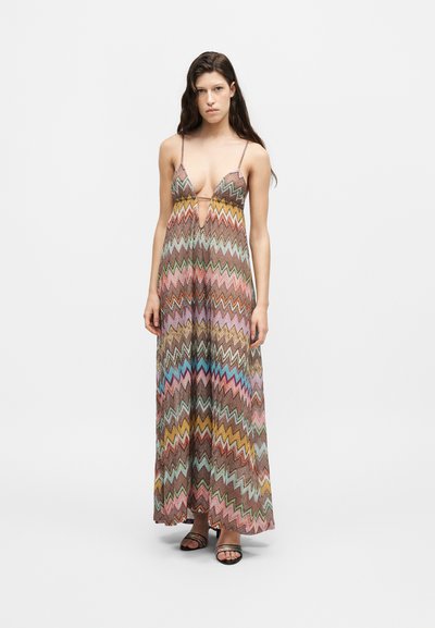 Femme portant une robe longue sans manches à motif zigzag multicolore avec bretelles fines et sandales noires ouvertes à l’avant, debout sur un fond uni.