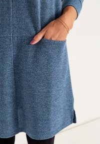 Rippstrickkleid in sanftem Blau, mit einer Fronttasche und seitlichen Schlitzen, langen Ärmeln und lockerer Passform. Sichtbares Detail des strukturierten Stoffes.