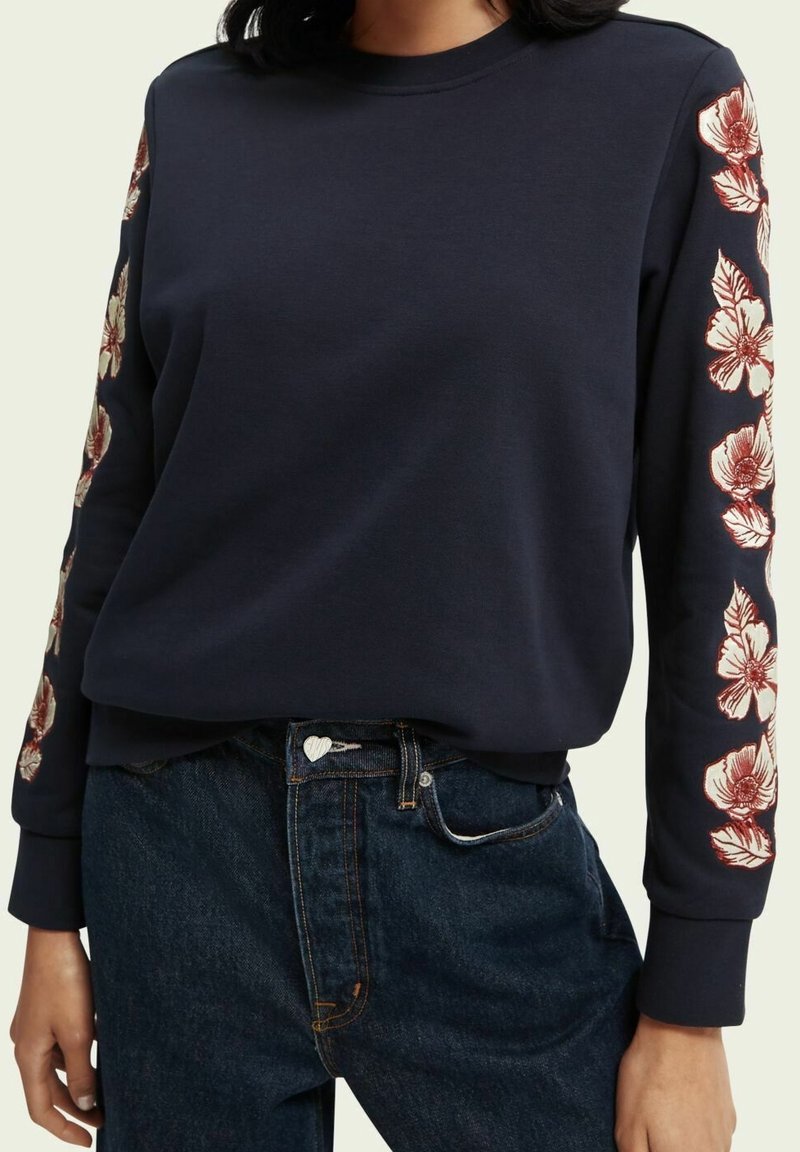 Femme portant un sweat-shirt bleu marine avec des motifs floraux rouges et blancs le long des deux manches, associé à un jean bleu foncé avec un bouton en forme de cœur.