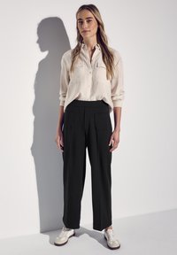 Beige košile na knoflíky s dvěma náprsními kapsami, kombinována s volnými černými kalhotami a béžovými teniskami, stojící na neutrálním pozadí.