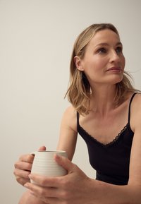 Camisola negra con detalles de encaje, sosteniendo una taza de cerámica gris claro con textura. Cabello corto y ondulado, fondo neutro. Enfoque en los detalles del producto.