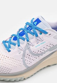 Chaussure de trail Nike de couleur rose clair et gris, dotée d'un mesh respirant, de lacets bleus, d'une texture perforée et d'une semelle beige robuste.