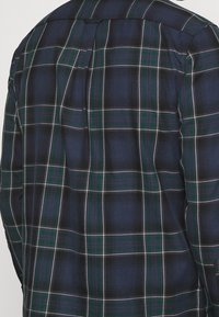 Vista trasera de una persona que lleva una camisa de cuadros en azul oscuro, verde y blanco de manga larga con yugo y pliegue central en la parte trasera.