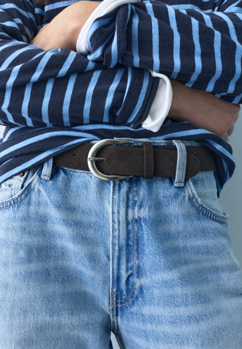 Personne portant une chemise à manches longues rayée bleue rentrée dans un jean en denim bleu clair avec une ceinture marron foncé, bras croisés sur la poitrine.