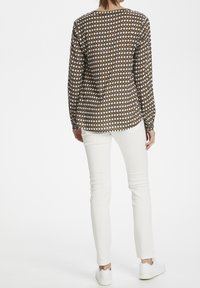 Femme portant un chemisier à manches longues à motif losanges marron et blanc, avec un pantalon blanc et des baskets blanches, se tenant de dos.