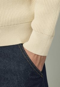 Des poignets de pull côtelés de couleur crème contrastent avec un jean en denim foncé, mettant en avant une coupe ajustée et un tissu tricoté texturé.