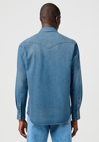Camisa de denim en azul claro con un canesú puntiagudo, mangas largas y puños con botones a presión. Textura suave con un diseño casual.