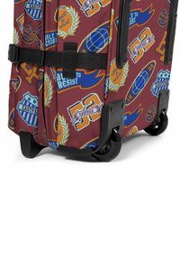 Valigia a rotelle in tessuto bordeaux, caratterizzata da vari loghi e design stampati in blu, giallo e bianco. Accenti neri sulle ruote e sul manico.