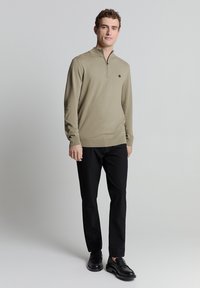 Beige zip-neck trui met lange mouwen, een gladde textuur, een geribbelde zoom, gedragen met zwarte broeken en zwarte loafers, staande tegen een lichte achtergrond.