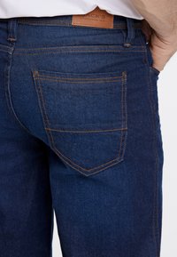 Jeans de mezclilla azul oscuro con costuras naranjas, que cuentan con un bolsillo trasero y una etiqueta de cuero con la marca. Textura suave, diseño ajustado.