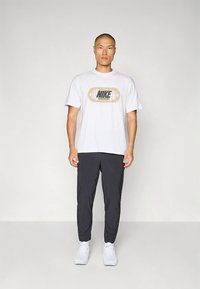 Valge puuvillane T-särk, mille keskel on halli ja oliivrohelise Nike logo, kombineeritud tumeda halli spordipüksidega ja valgete tossudega.