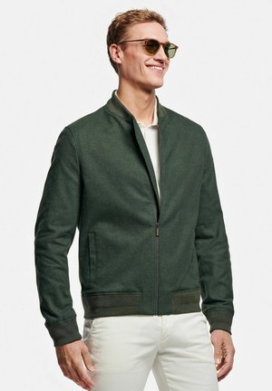 Lachende man met bruine zonnebril, donker groene jas met rits en witte broek, staand tegen een effen witte achtergrond.