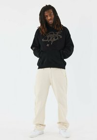 Sudadera negra con capucha y un diseño gráfico, combinada con pantalones beige claros y zapatillas blancas. Conjunto simple y casual con un corte holgado.