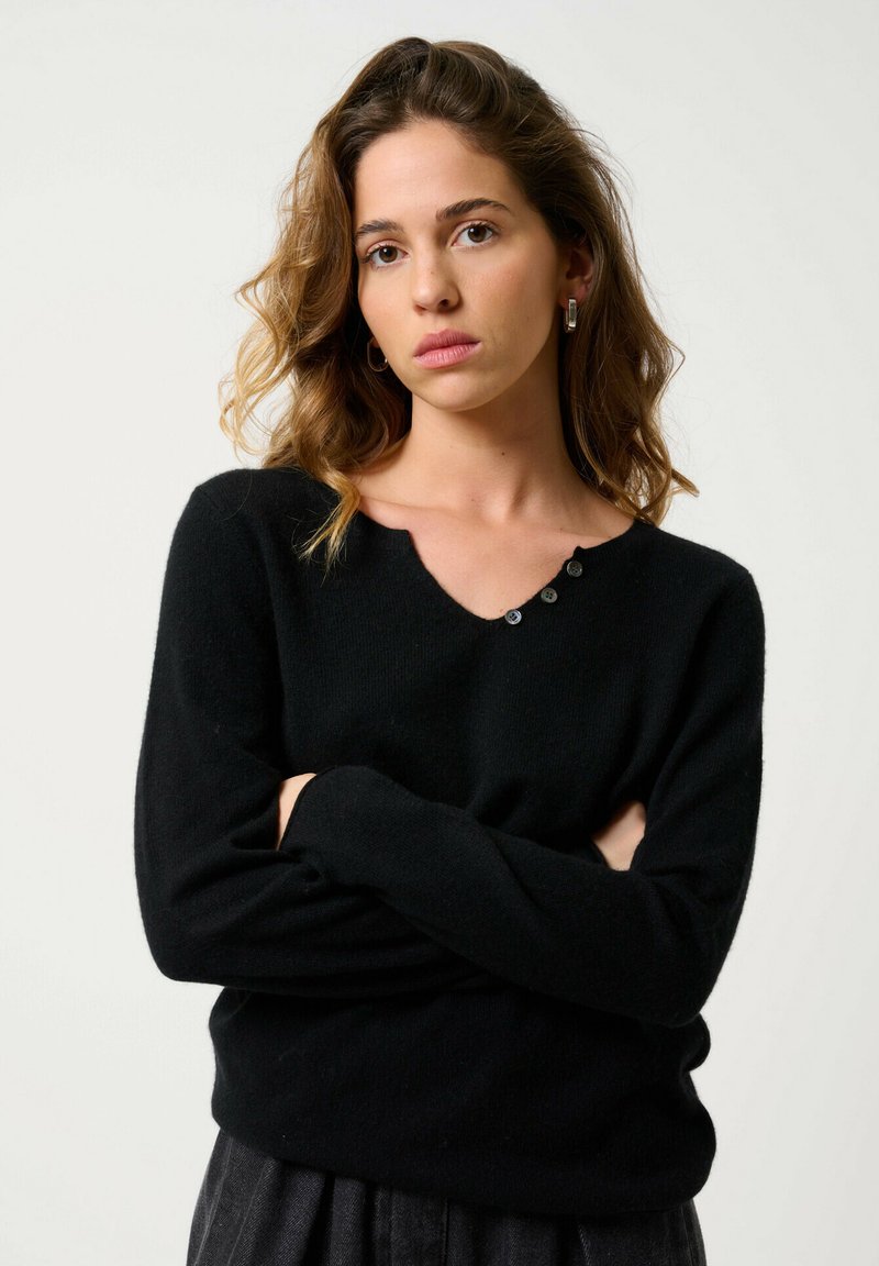 Pull noir avec un col rond, texture douce, manches longues et quatre boutons décoratifs sur l'épaule gauche.