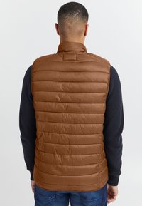 Braune isolierte Weste mit horizontalem Steppmuster, hohem Kragen und glatter Textur. Wird über einem dunklen Pullover getragen, Rückansicht gezeigt.