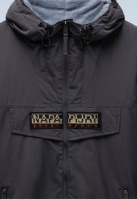 Veste à capuche gris foncé avec fermeture éclair centrale et un patch beige et noir indiquant "NAPAPIJRI geographic" sur la poitrine.