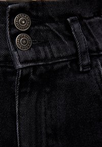 Les jeans en denim noir présentent une vue rapprochée de deux boutons métalliques avec logo, un tissu texturé et une ceinture cousue.