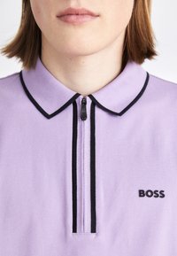 Světle fialová polo košile s černými akcenty, má zipový přední díl a vyšívané logo "BOSS" na levé straně. Hladká textura.