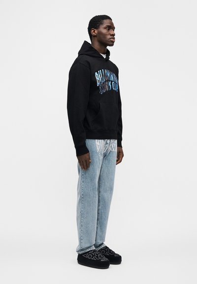 Μαύρο φούτερ με κουκούλα και μπροστινή τσέπη, με έντονο μπλε και λευκό λογότυπο. Ανοιχτό μπλε τζιν με σχέδιο παϊένσλι. Μαύρα παπούτσια με διακοσμητικές λεπτομέρειες.