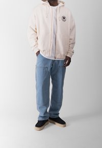 Sudadera cremosa con cremallera y cordones, llevada sobre una camiseta blanca; combinada con unos vaqueros de mezclilla holgados en azul claro y zapatos de ante negros.