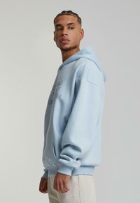 Gaiši zils hoody, kas izgatavots no mīkstas auduma, ar priekšējo kabatu, ripinātām aprocēm un apakšmalu, ar dekoratīvu tekstu kontrastējošā krāsā.